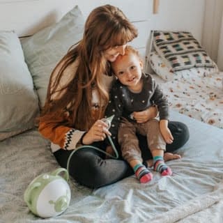 Mama siedzi z uśmiechniętą córką na łóżku, trzymają elektryczny aspirator do nosa Nosiboo Pro