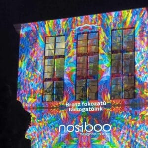 Logo Nosiboo wyświetlone na kolorowym światłem na budynku, informujące o brązowym poziomie sponsoringu Festiwalu Światła Zsolnay w Pécs.