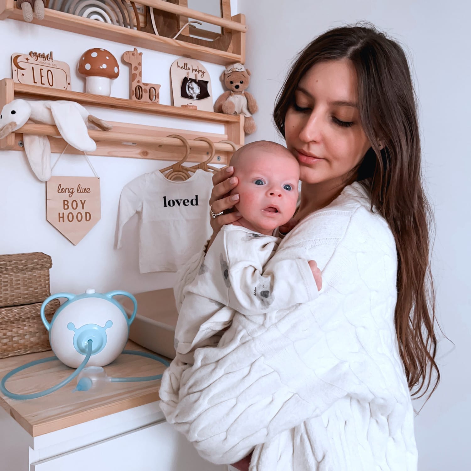 Ładna mama trzymająca w ramionach swoje urocze nowonarodzone dziecko, stojąca w pokoju dziecięcym obok elektrycznego aspiratora do nosa Nosiboo Pro.