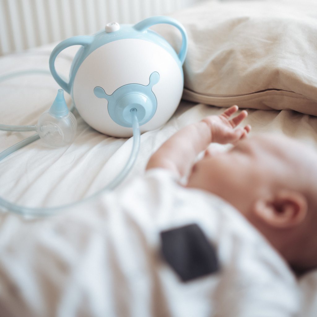 Chłopiec leży w łóżku, obok niego niebieski elektryczny aspirator do nosa Nosiboo Pro