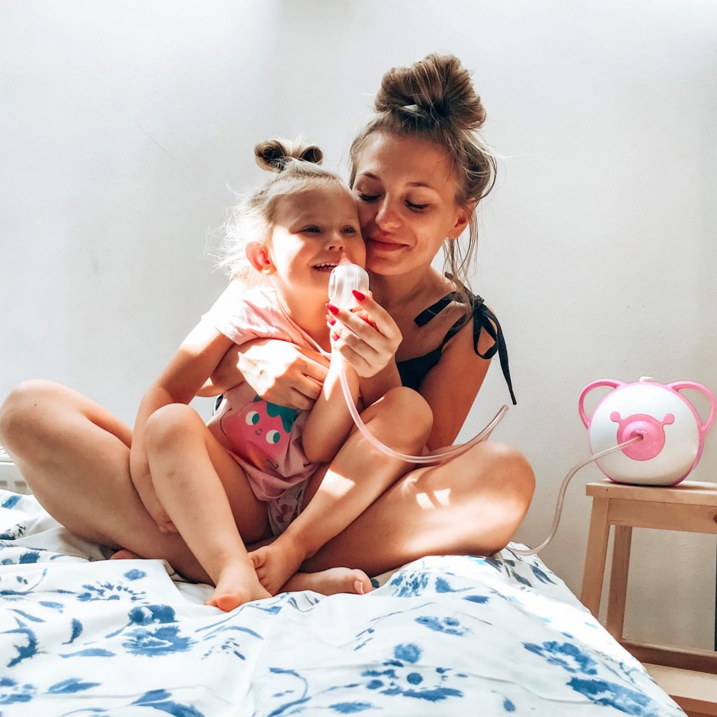 Uśmiechnięta mama i jej córka siedzą na łóżku, używają elektrycznego aspiratora do nosa Nosiboo Pro