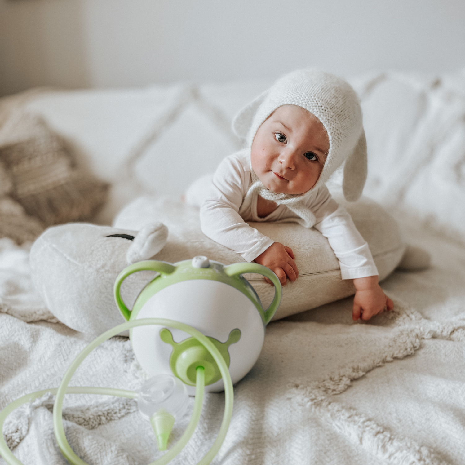 A baby in a cute beanie on a bed next to the Nosiboo Pro Electric Nasal Aspirator for cleaning little noses.
